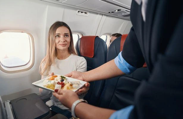 Пассажирка, сидя у иллюминатора в самолете, получает еду от бортпроводницы.